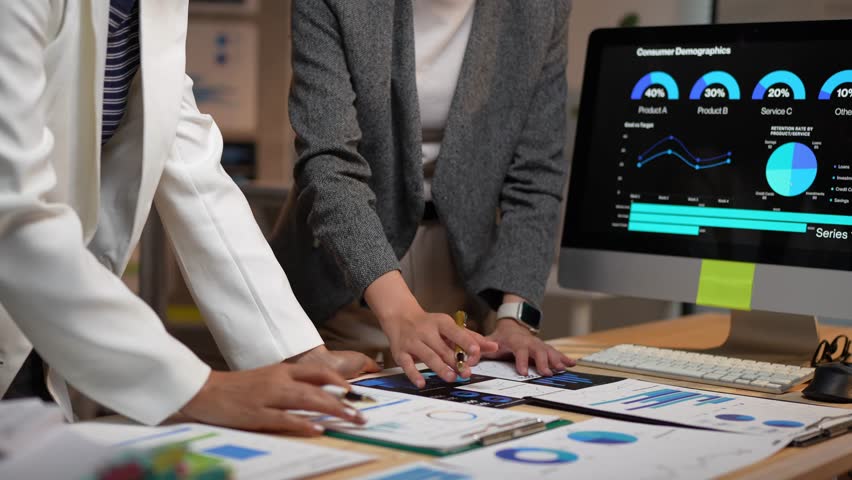 Two businesswomen collaborate, analyzing consumer demographics displayed on a computer screen and printed charts, pointing and discussing data insights to optimize marketing strategies - Powered by Shutterstock - Get 15% off with code: PIKWIZARD15