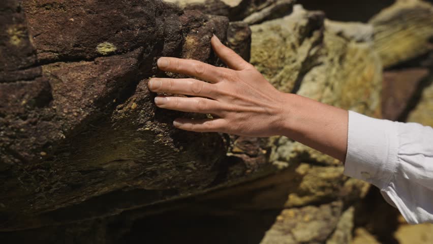 A female hand glides over textured sandstone rocks, capturing the rough tactile sensation and geological volume of the material.