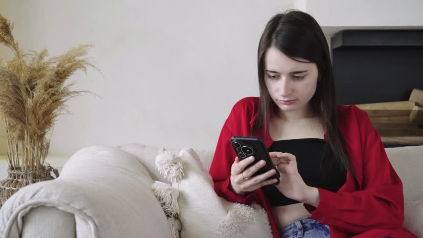 A young brunette woman sits at home on the couch, browses social media on her smartphone and finds bad news that upsets her, she starts crying