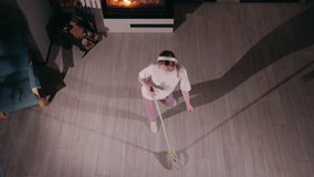 Young woman dancing with a mop while cleaning, viewed from above. She wears a white t-shirt, pink sweatpants, and white socks, listening to music on headphones and making household chores fun in a - Powered by Shutterstock - Get 15% off with code: PIKWIZARD15