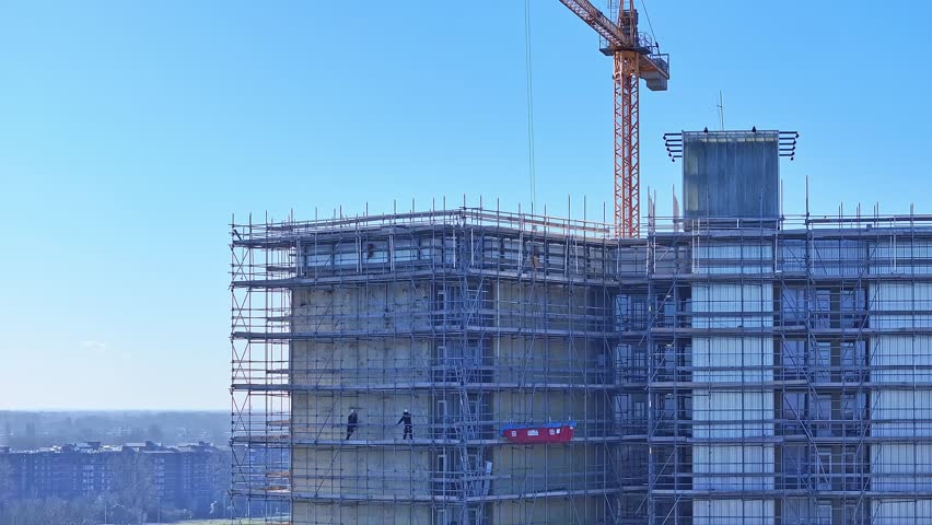 Crane On Top Of Concrete Building, Construction Site With Scaffolds and Construction Workers, Aerial View