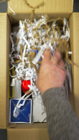 Vertical video - Closeup overhead shot of a man’s hands adding shredded paper and card over shipping items in a cardboard box, then adding a padded bag on top to complete the packing. 