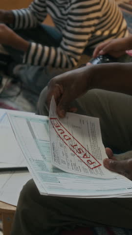Vertical shot of unrecognizable Black man looking through past due bills and eviction notice, sitting on couch