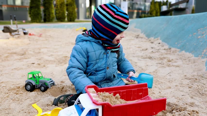 Young child plays with toy trucks in sandy playground on a cool day wearing warm clothing