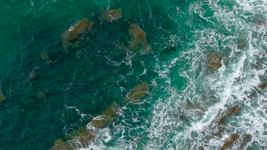 Ocean blue waves break on high cliff of rocky mountain. Turquoise sea crashing against steep stone wall from above. Powerful surf slamming jagged coastal rock face. Foamy water hitting vertical cliffs