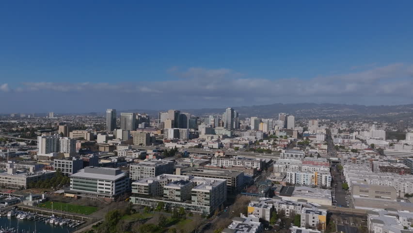 Aerial footage of downtown Oakland, California, showcasing its mix of modern high-rises, dense urban neighborhoods, and a backdrop of rolling hills under a clear blue sky.