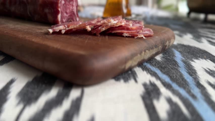 A raw-dried Matykova sausage, similar to a robust prosciutto or other cured ham, served sliced on a wooden board next to Turkish-style tea glasses on an ikat placemat.