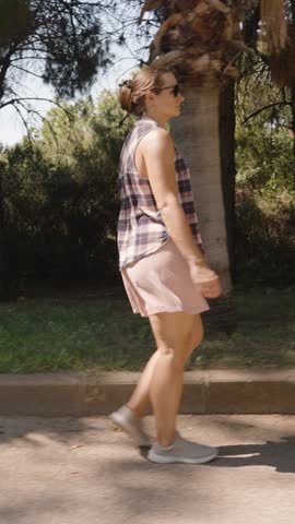 Vertical video. Woman walks past a Bench under the Shade of Trees along a Quiet Park Path, her steps soft on the pavement.