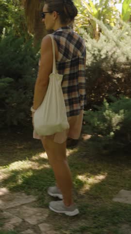 Vertical video. Woman walks on a Path through a Sunlit Garden, surrounded by Lush Tropical Plants and casting Soft Shadows on the Grass.