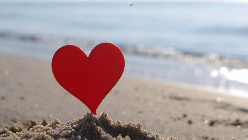 Plastic stick shape of red heart in sand beach of sea shore on background of sea waves on sunny summer day. Concept Love relationship romance amorousness amour St Valentines Day
