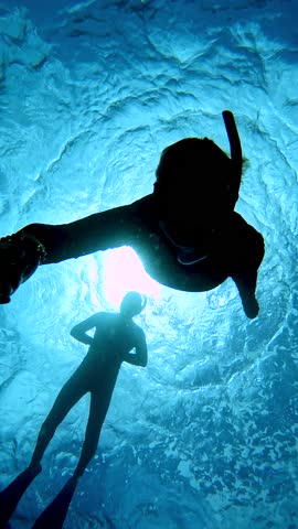 Silhouette of two freedivers floating through sunbeams in Maldives. An ocean Water surface with bright sun underwater behind ripples.
