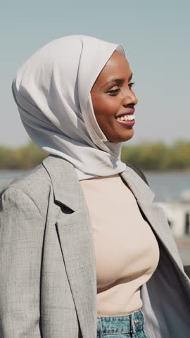Smiling Arabic woman with hijab walks along embankment past river boat station slow motion. Black lady in city in sunny weather. Happy Muslimah at promenade