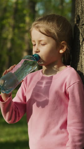 Tired little girl drinks water leaning on old tree trunk in green park closeup. Stay hydrated to avoid heat stroke. Refreshing drink in hot weather