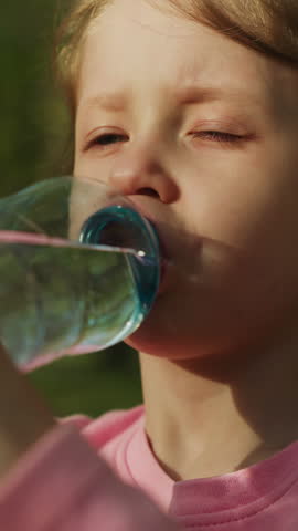 Thirsty little child drinks clear fresh water by old tree trunk in city park closeup. Girl refreshes after active playing. Heat stroke prevention