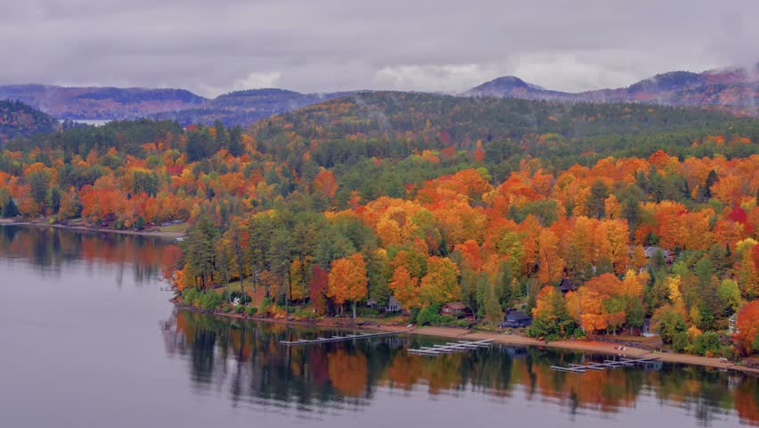 A breathtaking drone view of an Adirondack lake framed by vibrant fall foliage and drifting fog, with mountains rising in the distance during peak autumn season.