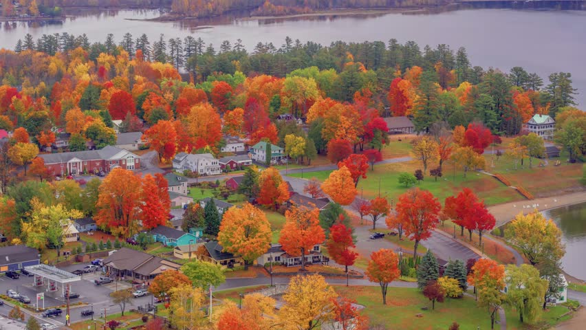 Charming drone scene of a rustic town at the edge of an Adirondack lake, wrapped in autumn colors and mist with a scenic mountain backdrop in fall season.