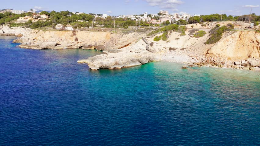 A coastal cityscape with a rocky coastline and some vegetation. A road runs along the coast with cars visible. There are buildings and a docked ship in the background under a sky with scattered cloud
