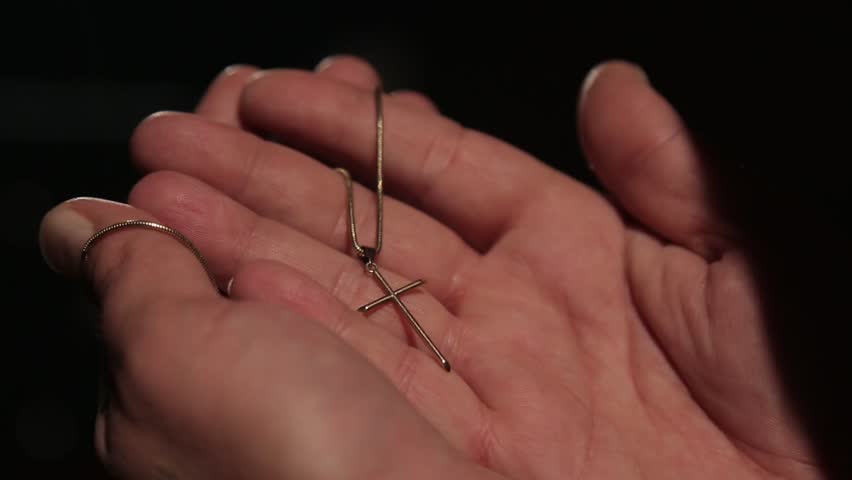 A silver cross necklace rests on a dark, striped wooden table. A hand enters the frame, touches the necklace briefly, and then leaves, leaving the cross behind.