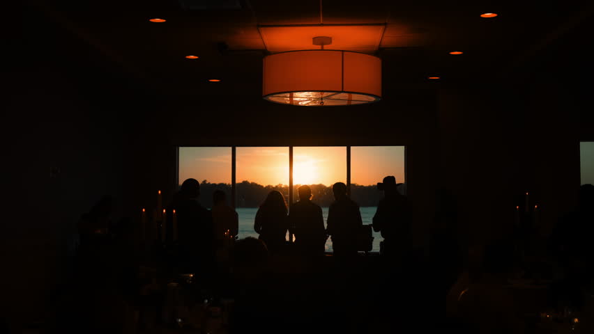 Soft unrecognizable silhouettes of random people in golden sunset light streaming through window, camera view from the inside of venue