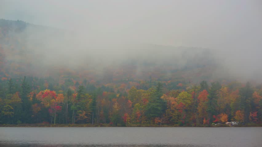 This beautiful footage captures Adirondack Park in New York during the fall season, showcasing a foggy lake surrounded by forests adorned with vibrant autumn foliage.