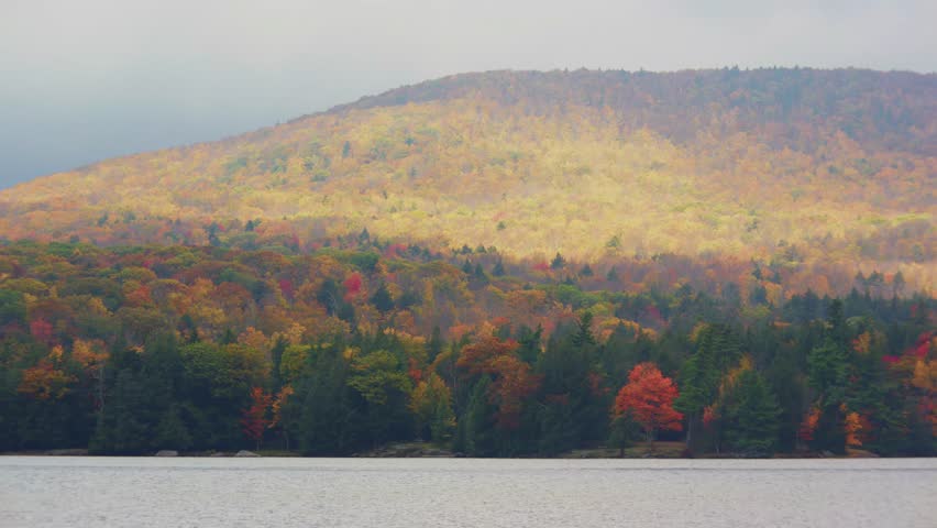 This stunning footage captures the beauty of Adirondack Park in New York during the fall season, with a foggy atmosphere over a serene lake surrounded by vibrant autumn foliage.