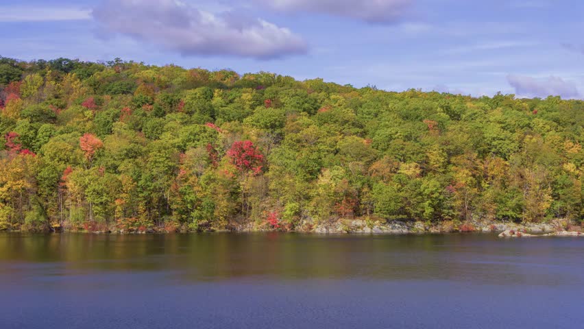 This 4K drone footage of Canopus Lake in Kent, NY, captured during autumn, highlights the lake surrounded by vibrant fall colors and soft cloudy skies.