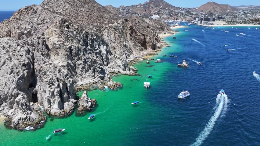flying along the rocky coastline above turquoise water near Pelican Rock in Cabo Bay near Cabo San Lucas marina with many tourist boats driving around and people swimming in the bright blue water 