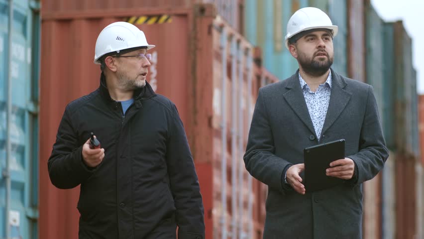 Fellow engineers walking and discussing in front of a stacked cargo container. A shipping manager and a loading dock worker work in the commercial dock industry. Logistics transport business.