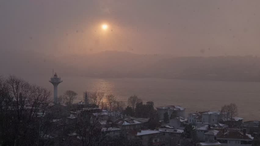 A stunning view of the Bosphorus during a red sunset, with snow gently falling over the scene. The contrast of snow and sunlight creates a magical atmosphere, as ferries cruise along the water.