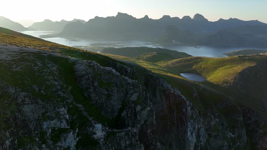 A drone view of Ryten, Lofoten Islands, Norway, showcasing rocky hills covered in lush green, with the distant sea surrounded by towering hills