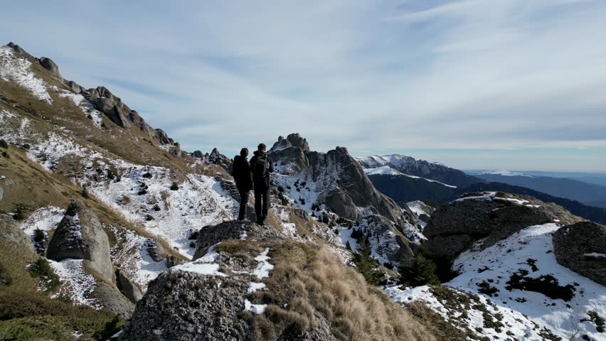 A drone view of two people standing on top and looking at the Ciucas mountain peaks what covered in snow on a sunny day in Romania