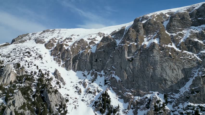 A drone view of the snow-covered Bucegi mountains of Romania during winter with rocky peaks and clear blue sky in Brasov
