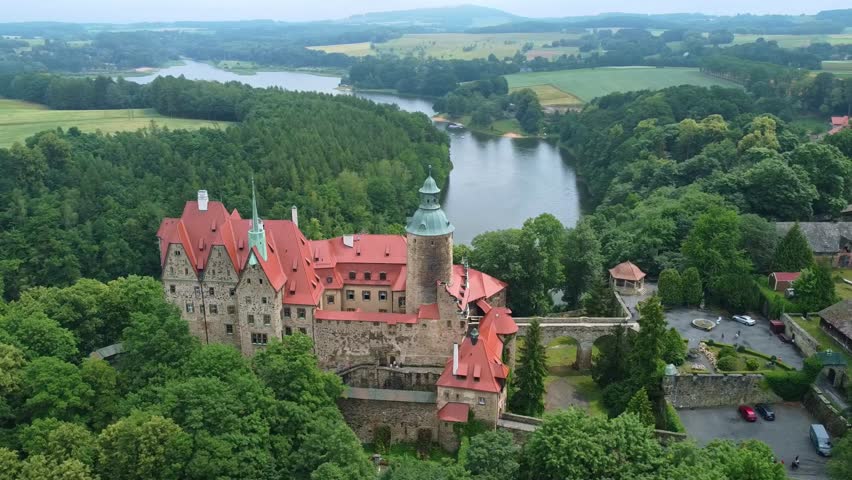 Flyby shot of medieval Czocha Castle perched on water’s edge in scenic green forest, Poland
