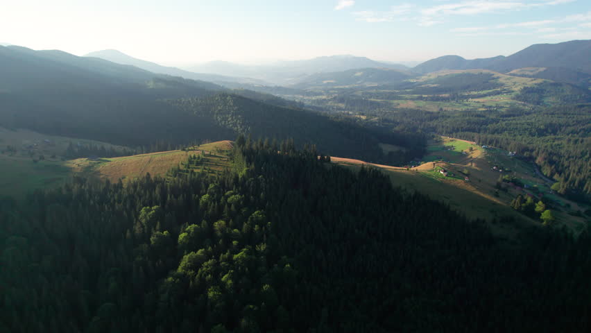 Aerial view of lush valley with scattered houses and farms, surrounded by dense forests and rolling hills. Morning sunlight casts long shadows, creating picturesque landscape.