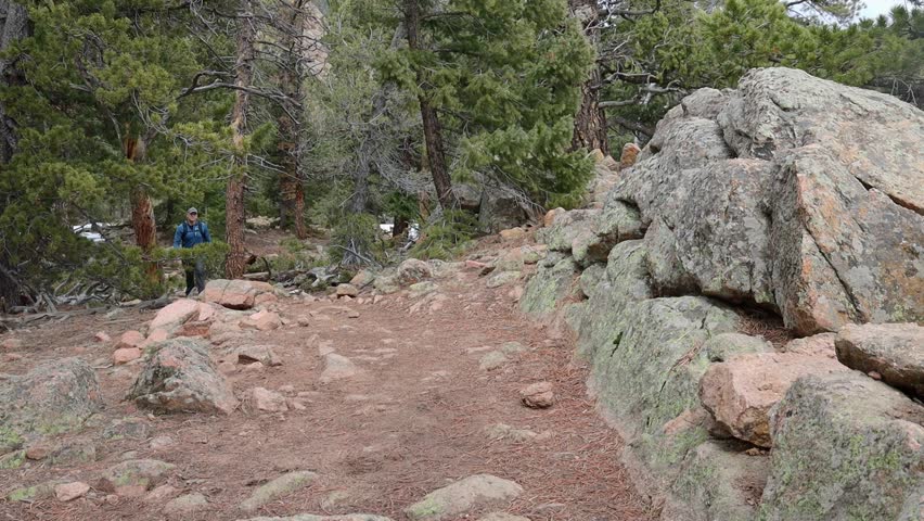 One male hiker with a backpack walking up a steep trail and by a rock wall with snow on the ground. Filmed in Staunton State Park during the spring.