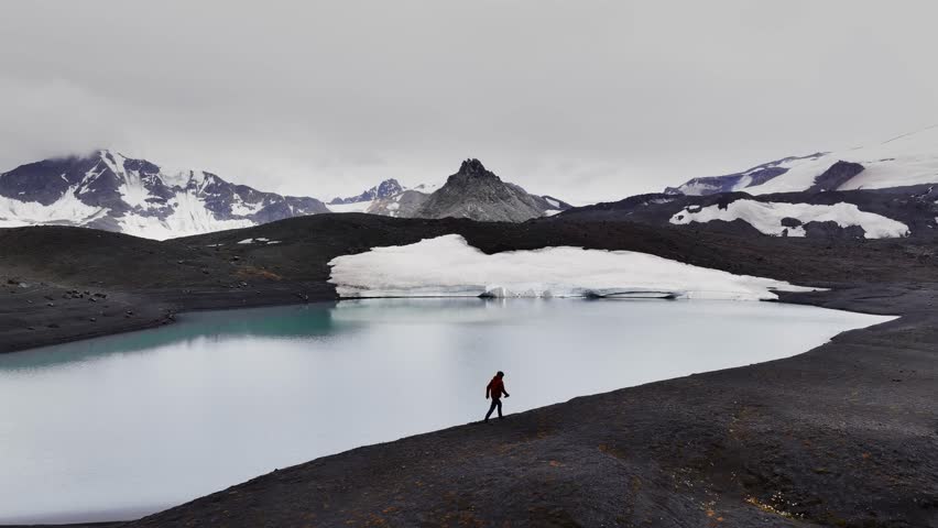 A man walks through volcanic rocks surrounded by a mountain lake against the backdrop of epic mountains and cliffs of the volcanic terrain with glaciers