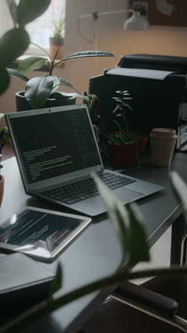 Vertical shot of laptop with code on screen, digital tablet and printer on desk decorated with potted plants in cozy modern office