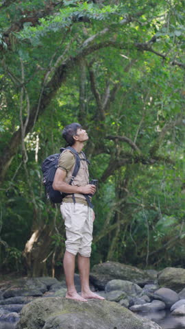 An Asian male traveler stands barefoot on a rock in the forest, birdwatching through binoculars, embracing eco-tourism and a deep connection with nature