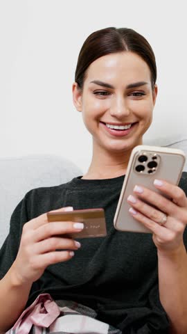 Vertical video. A woman makes an online payment or purchase from the comfort of her home, using her credit card and smartphone. The concept of convenient and safe online shopping.