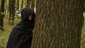 Painted hand, Shadowy figure, Mysterious figure. In shadowy forest, hooded figure with painted hand grips tree trunk, evoking secrecy and suspense. - Powered by Shutterstock - Get 15% off with code: PIKWIZARD15