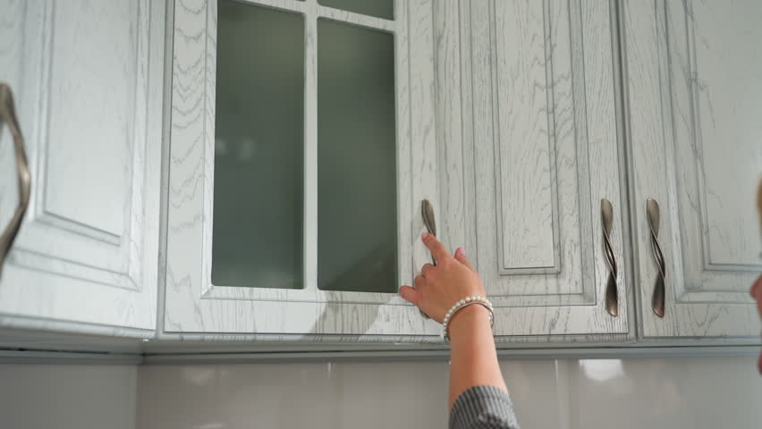Close up of young adult with beads adorned hand opening kitchen cabinet retrieving glass cup from upper shelf, captured against modern white cabinet background with soft natural lighting
