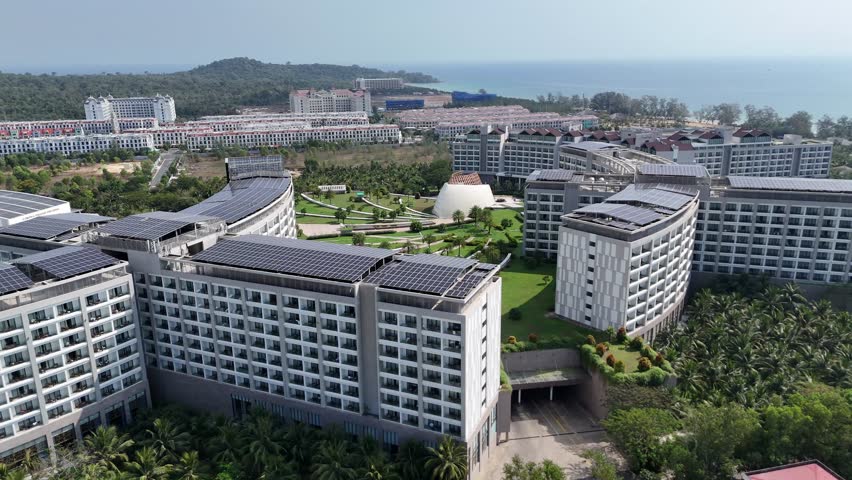 Afternoon aerial footage of a sprawling coastal resort with large modern buildings, lush greenery, a distinctive dome, and a sandy beach meeting the blue sea. Solar panels fixed in the buildings
