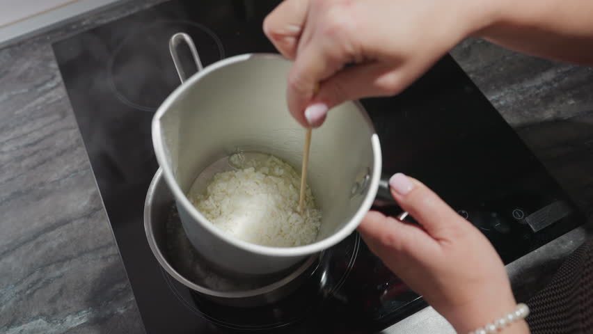 Female chandler melts tallow flakes inside metal mixing bowl placed in pot of hot water on electric stove, using double boiler method to safely liquefy wax substance for candle making in home kitchen