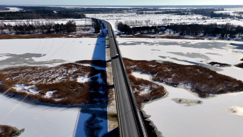 A road bridge cuts across icy marshland and a frozen river as cars move through a bright, snow-covered winter landscape under clear skies.