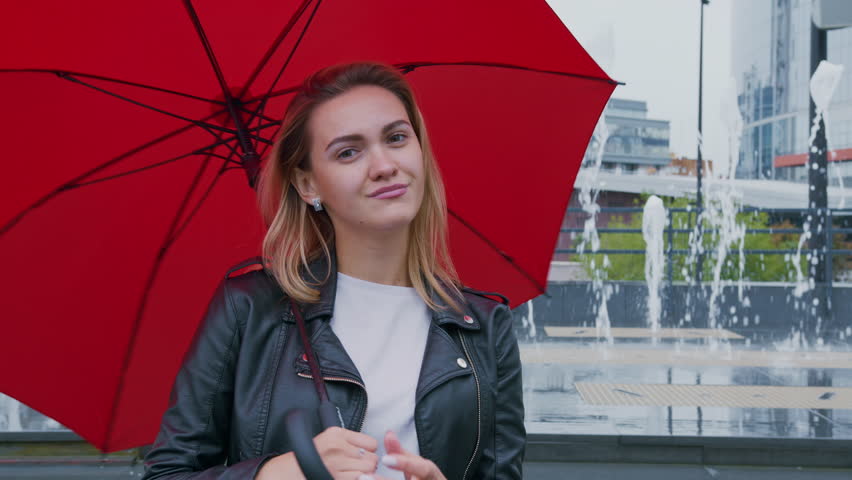 Young woman wearing red umbrella standing near city fountain, expressing various emotions while enjoying rainy urban environment with stylish confidence and cheerful demeanor