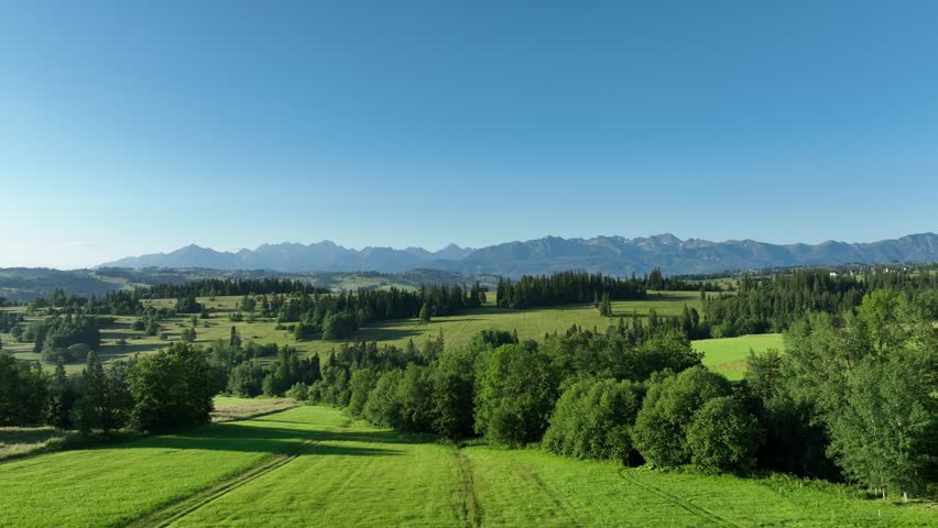 Aerial shots of a picturesque mountain landscape. Gentle, green hills stretch in the foreground, while high mountains rise majestically in the distance. The whole is complemented by a clear, blue sky