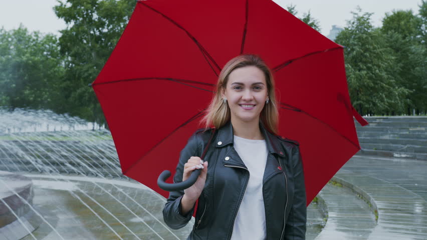 Blonde woman wearing leather jacket, standing near park fountain with red umbrella, radiating cheerful joy while smiling and looking at camera during rainy weather