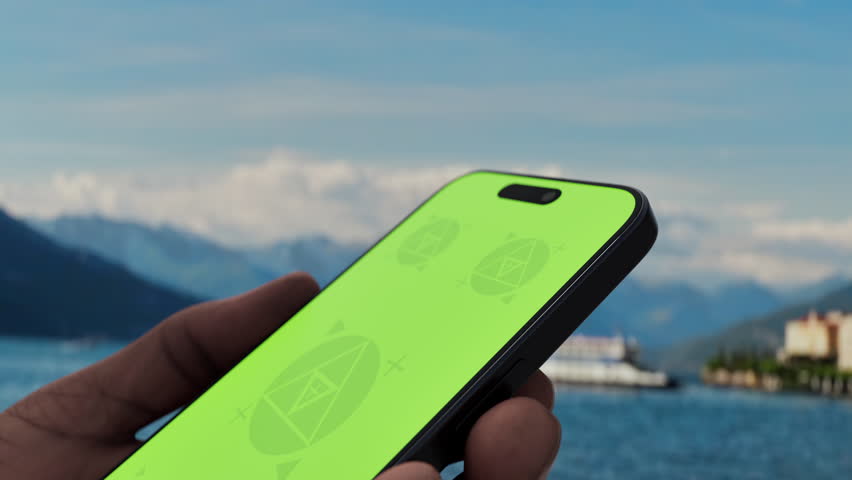 GREEN SCREEN CHROMA KEY A hand holding a smartphone with a green screen on Lake Como during the day, showing a beautiful scenic view. Ideal for travel or tourism apps