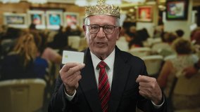 Senior man in suit holding card wearing crown in busy art gallery with blurred people and paintings in background, conveying a sense of authority and business in a cultured setting. - Powered by Shutterstock - Get 15% off with code: PIKWIZARD15