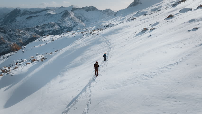 Two hikers trek through deep snow on a trail in the Pyrenees, surrounded by stunning snow-covered mountains and a clear sky during a winter day.